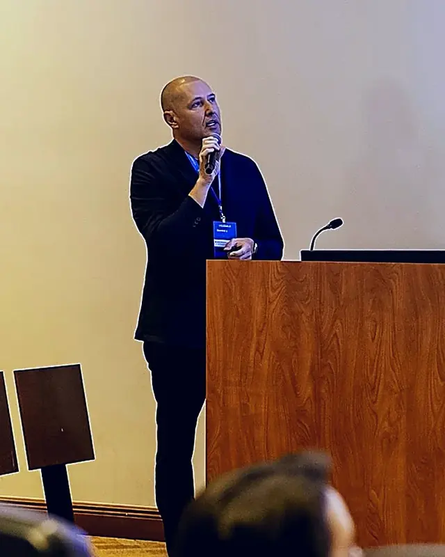 Dr. Igor Brazioli Slivinskis em pé ao lado de um púlpito de madeira, falando a um microfone de mão, com crachá no pescoço; em primeiro plano, a plateia fora de foco
