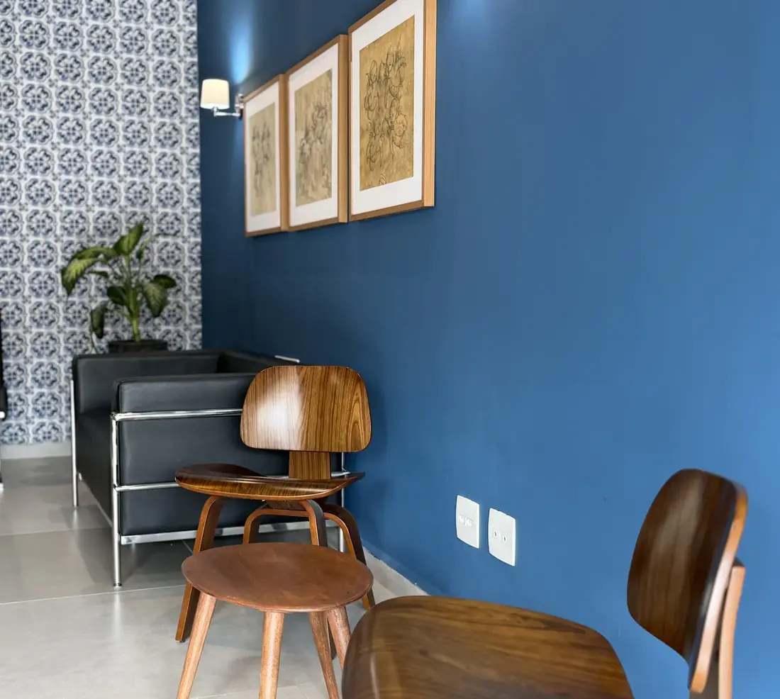 Recepção da clínica com parede azul, cadeiras de madeira, poltrona preta de couro e três quadros emoldurados.