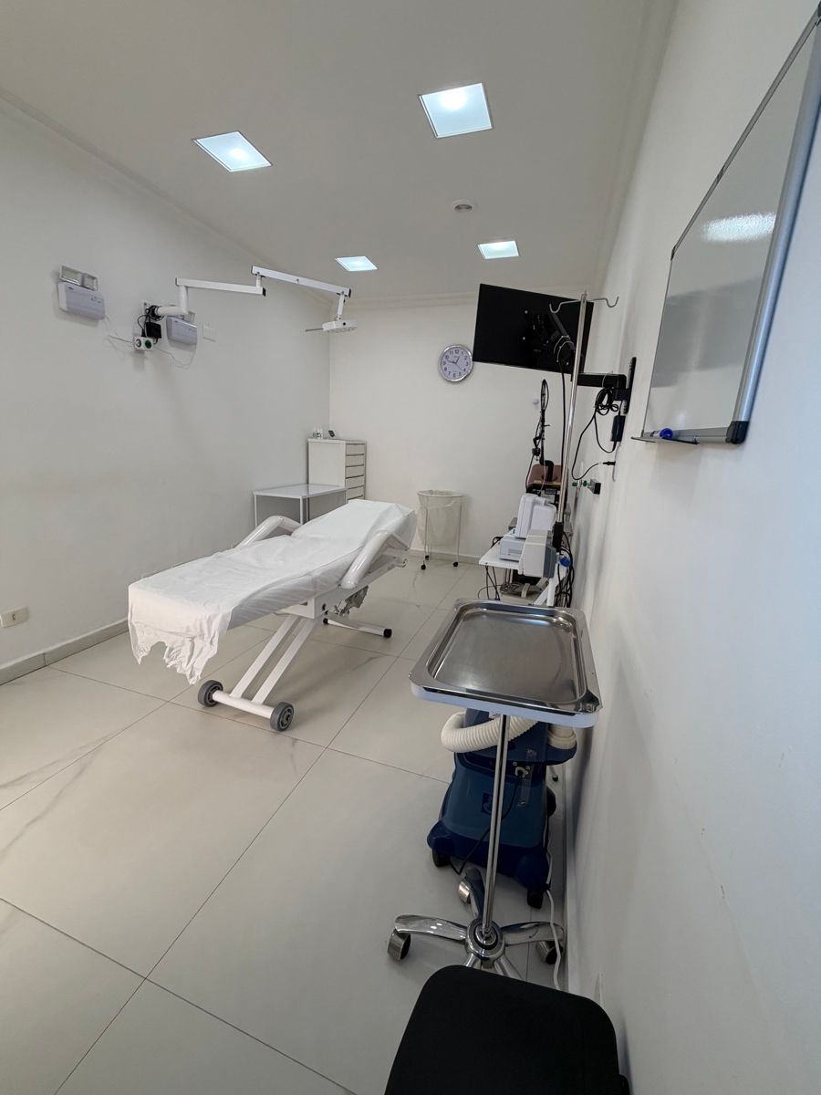 Sala branca com maca reclinável, foco de luz articulado fixado na parede, monitor de tela plana, suporte de soro, bandeja de inox sobre suporte com rodízios, equipamentos sobre carrinho e quadro branco.