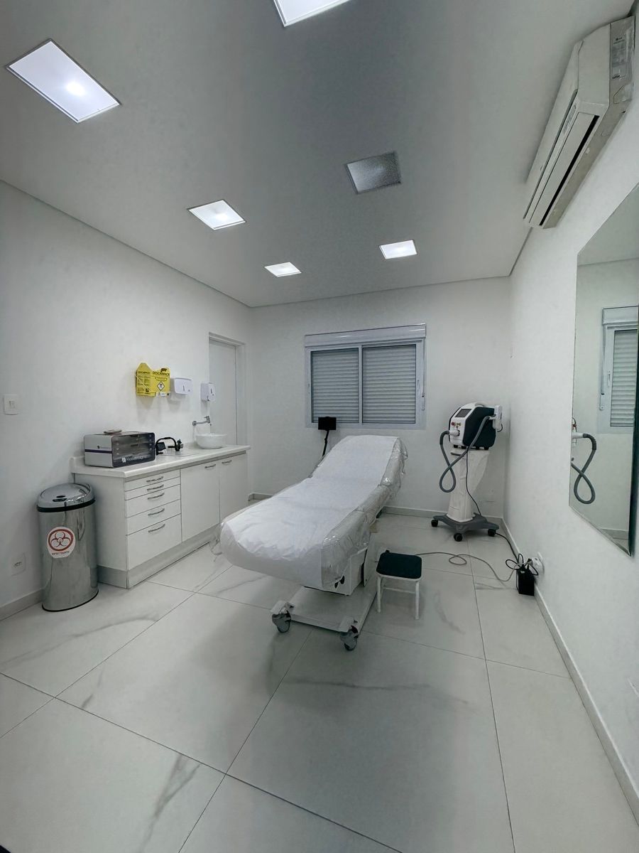 Sala de paredes brancas com maca revestida, bancada com pia e gavetas, equipamento sobre pedestal com rodízios, espelho na parede e aparelho de ar-condicionado.