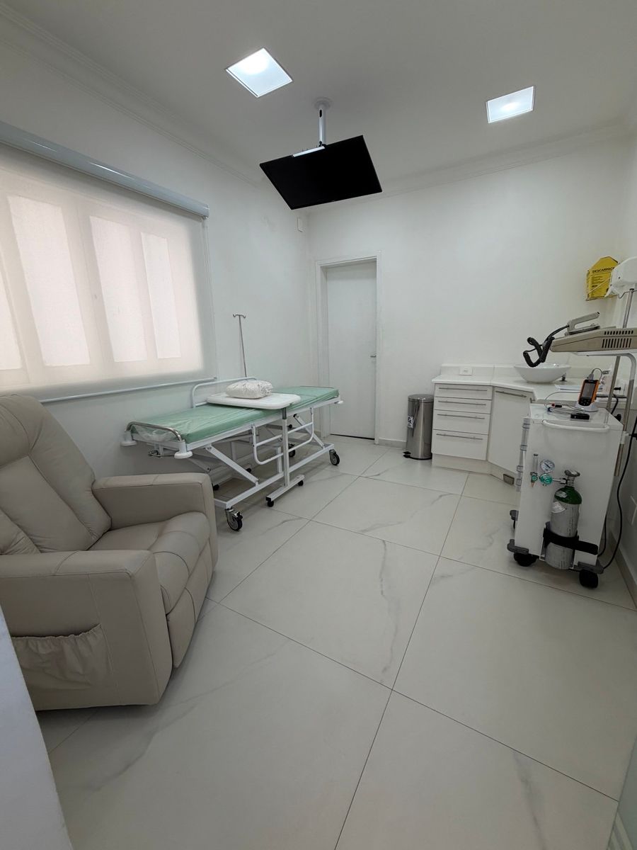 Sala branca com poltrona reclinável bege, maca com colchão verde e grades laterais, suporte de soro, televisor fixado no teto, bancada com pia e carrinho com cilindro de oxigênio.